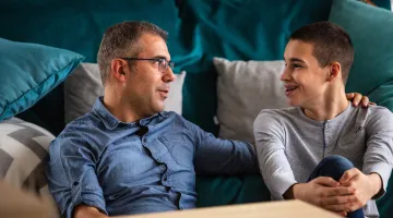 A father sits on the floor next to his teenage son, having a conversation with him.